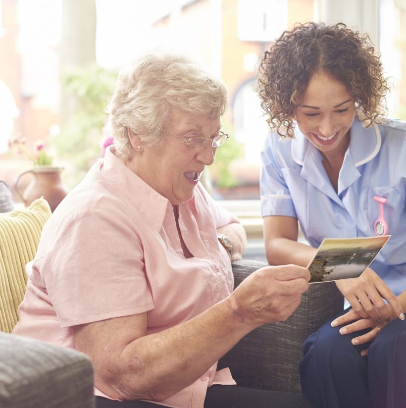 a senior woman in her home chatting with a friendly nurse who has called in to discuss her recovery .   they are sitting in a bay window of a house and the senior woman is showing a postcard from her grandchildren on holiday .